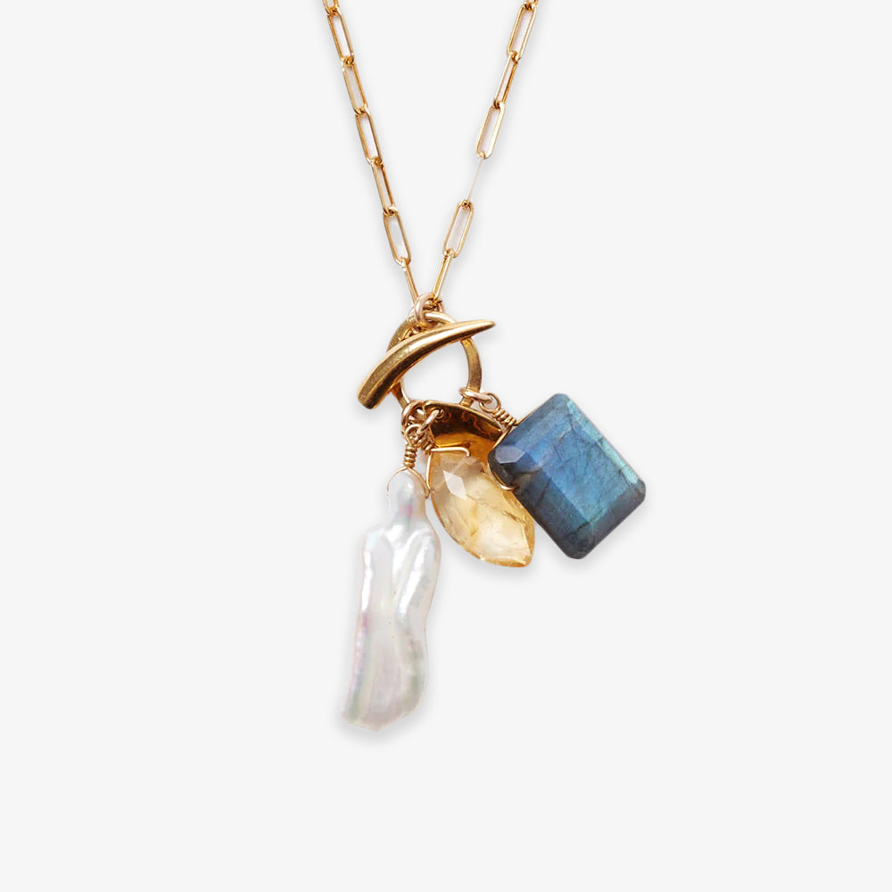 The Gemstone Mix Charm Necklace features a gold paperclip chain with three pendants—a white pearl, yellow gemstone, and blue rectangular stone—crafted as sustainable artisan jewelry.