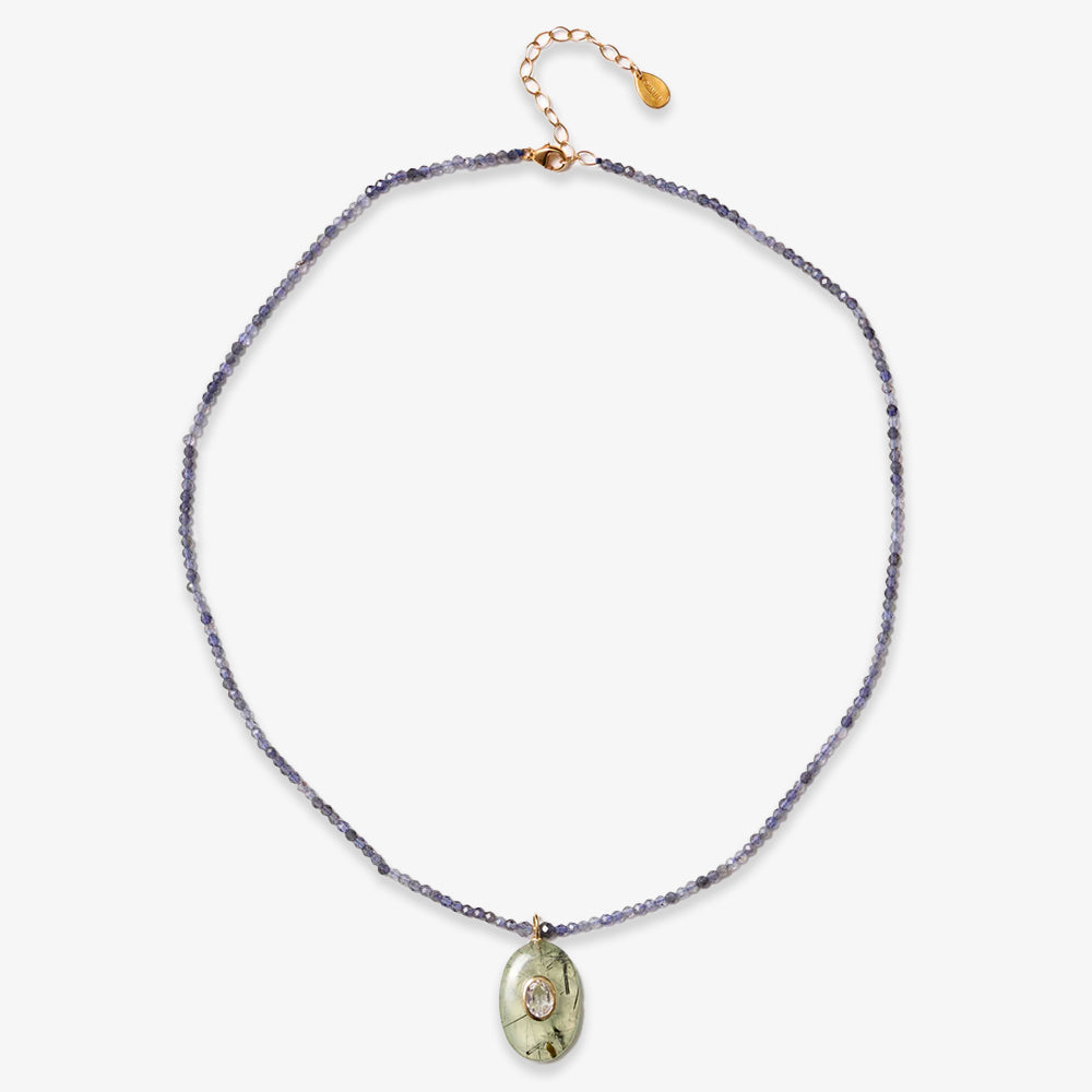 The Beaded Seed Gemstone Pendant Necklace features small blue beads, a gold clasp, and an engraved gold oval pendant with a clear crystal accent, highlighting artisan craftsmanship.