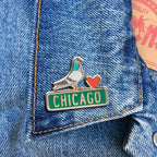 The Pigeon Pin, featuring a red heart and a green Chicago street sign, is pinned to a blue denim jacket, showcasing city pride and the Obama Foundation’s spirit.