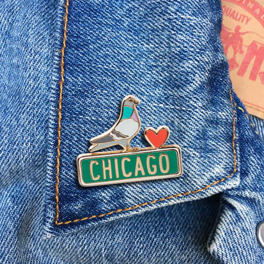 The Pigeon Pin, featuring a red heart and a green Chicago street sign, is pinned to a blue denim jacket, showcasing city pride and the Obama Foundation’s spirit.