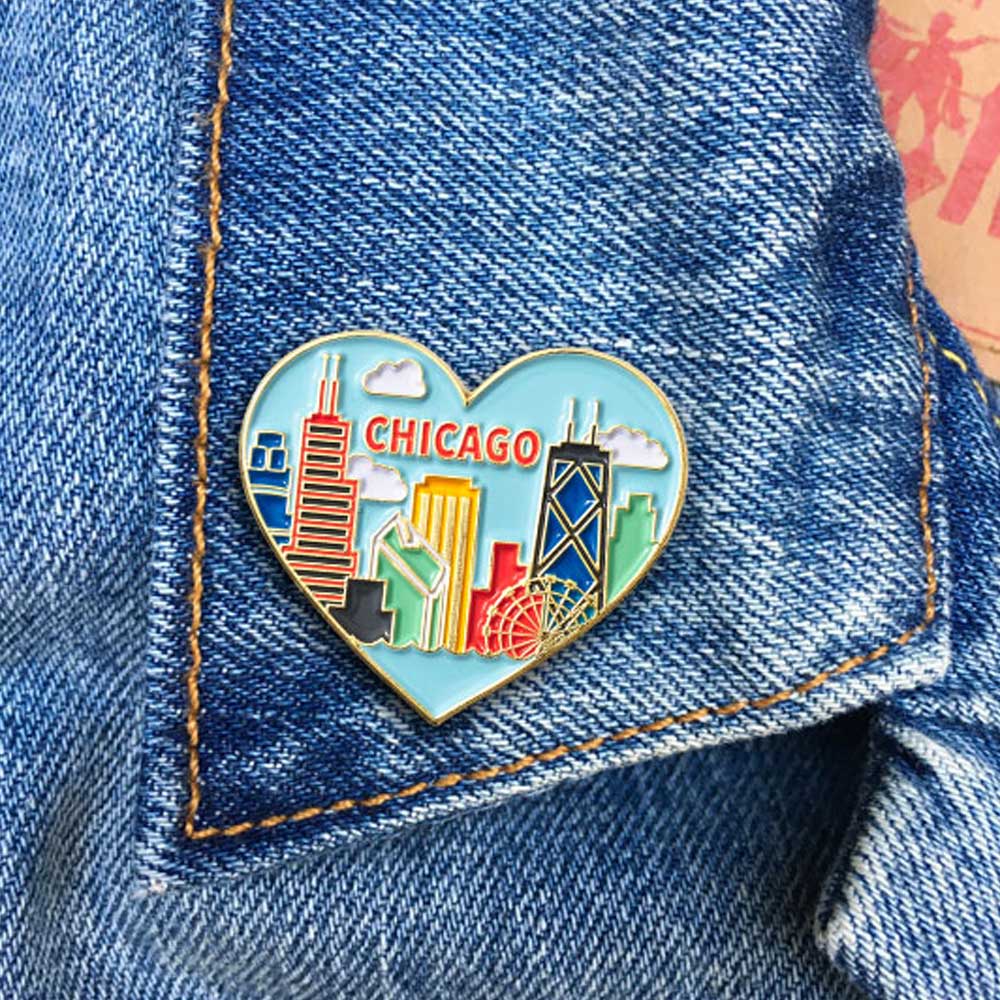 The Skyline Heart Pin is a heart-shaped enamel pin with a colorful Chicago skyline and "CHICAGO" text, shown on blue denim. It's the perfect accessory for city lovers and Obama Foundation fans.