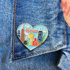 The Skyline Heart Pin is a heart-shaped enamel pin with a colorful Chicago skyline and "CHICAGO" text, shown on blue denim. It's the perfect accessory for city lovers and Obama Foundation fans.