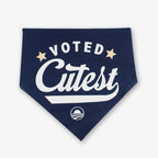 The Voted Cutest Kerchief is a navy dog bandana featuring bold white "Voted Cutest" text, two gold stars, and a small circular rising sun logo—an adorable accessory for your pup.