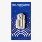The Obama Presidential Center Building Lapel Pin is a white handcrafted clay pin shaped like a building, presented on a blue card with white polka dots and text. Designed by an Obama Leader, it makes a unique architectural accessory.