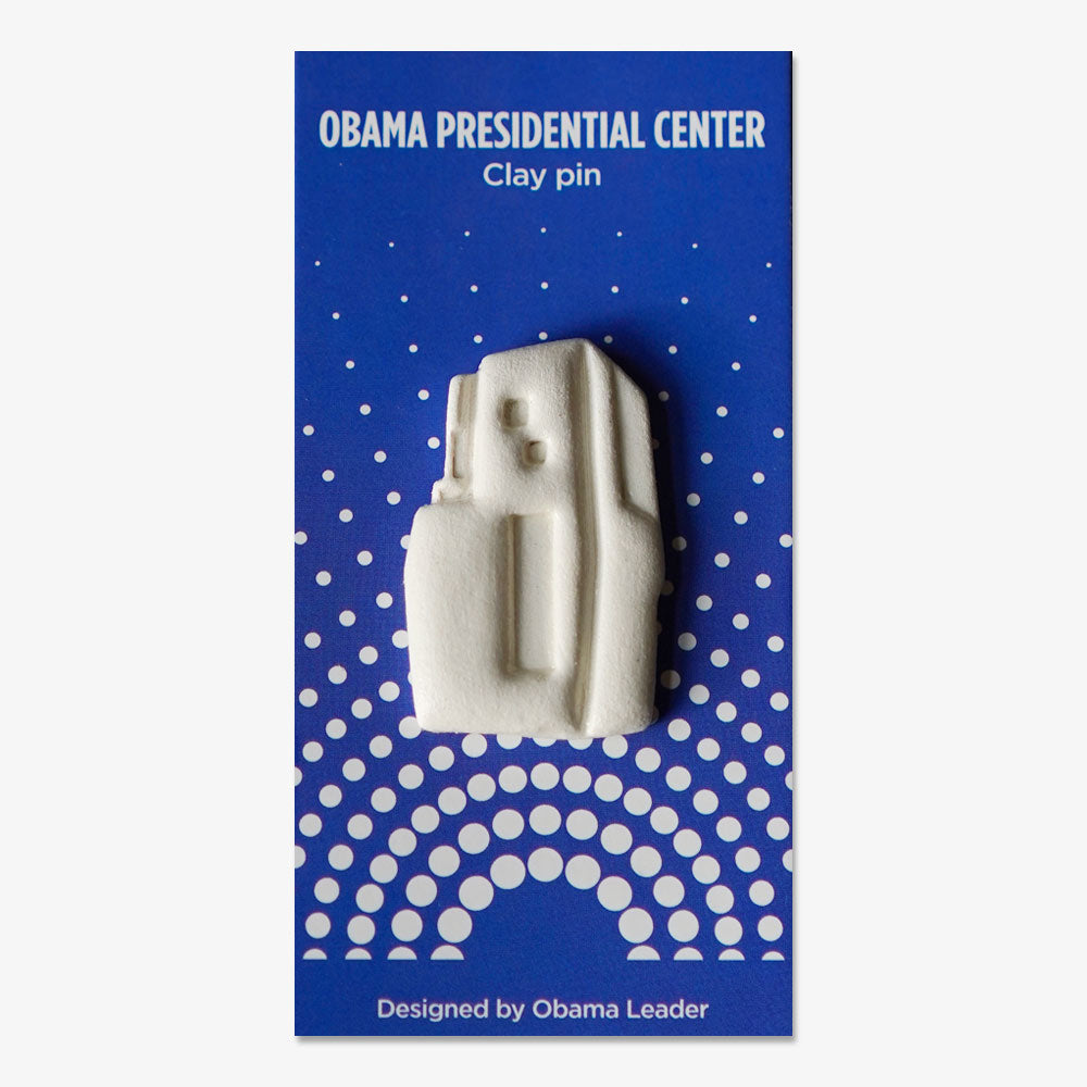 The Obama Presidential Center Building Lapel Pin is a white handcrafted clay pin shaped like a building, presented on a blue card with white polka dots and text. Designed by an Obama Leader, it makes a unique architectural accessory.