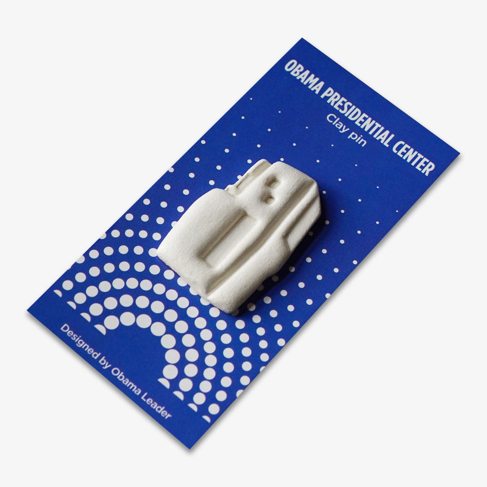 The Obama Presidential Center Building Lapel Pin is a handcrafted clay pin shaped like the center, attached to a blue card labeled "Obama Presidential Center" with dotted patterns, creating a distinctive architectural accessory.