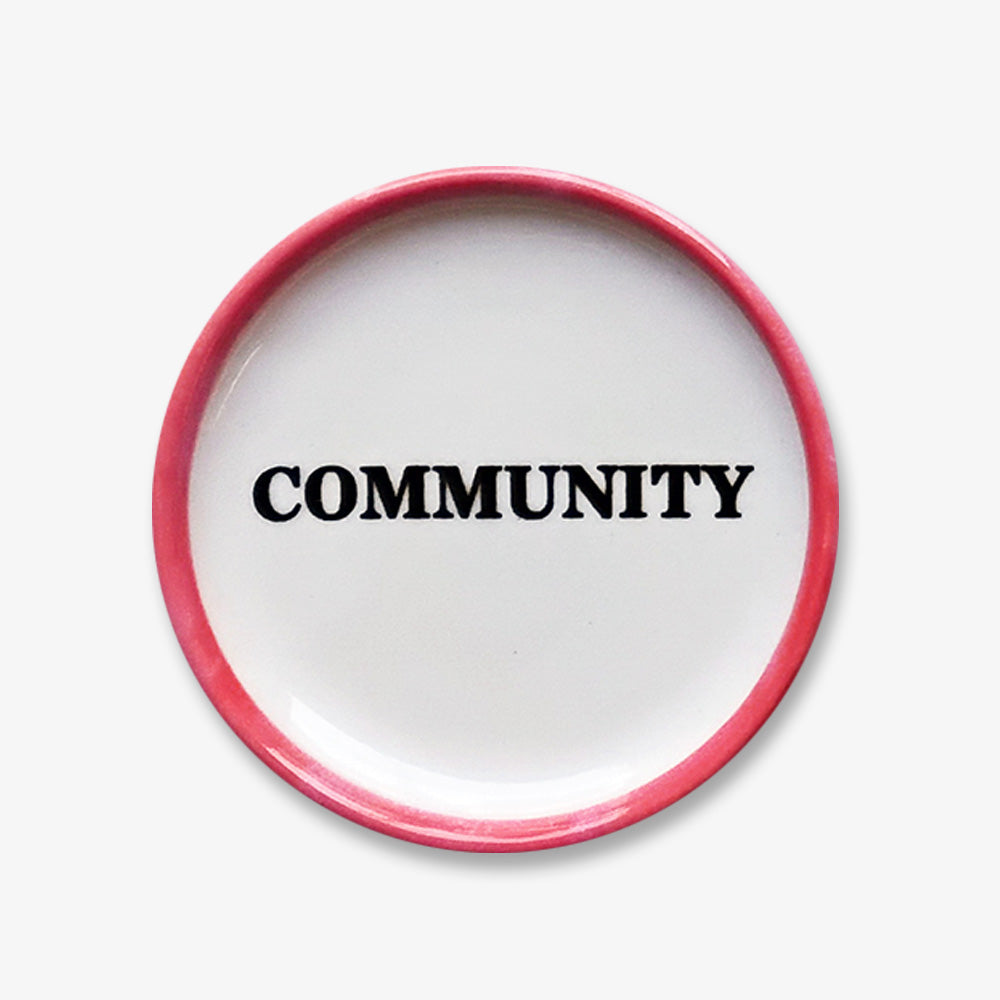 The Community Trinket Dish is a handmade porcelain piece featuring a pink rim and "COMMUNITY" in bold, black letters at its center. Perfect for small treasures, it stands out beautifully on any light surface.