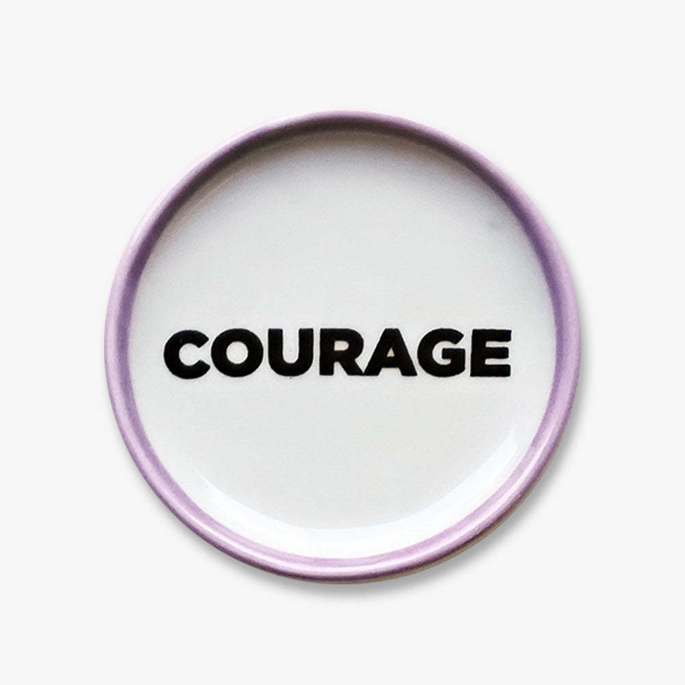 The Courage Trinket Dish is a small round handmade porcelain keepsake with a light purple rim and "COURAGE" in bold black letters at the center, inspired by the Obama Foundation.