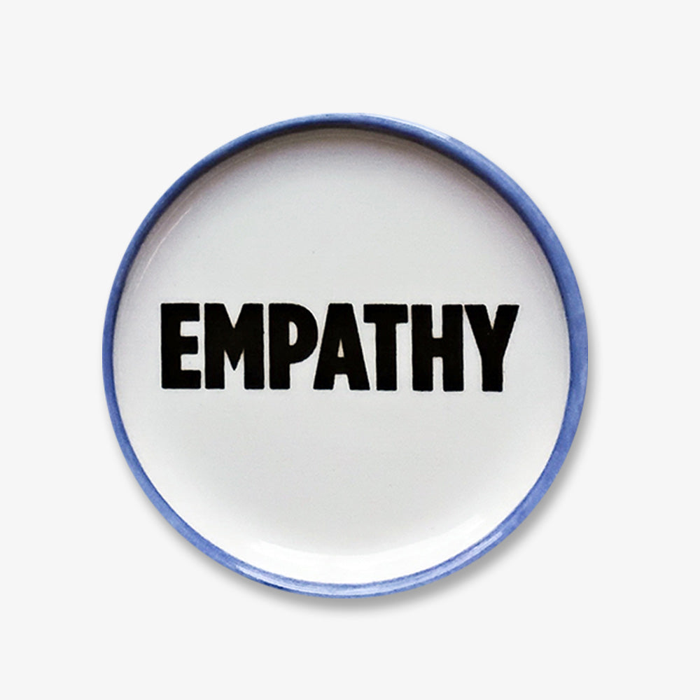 The Empathy Trinket Dish is a handmade porcelain dish with a blue rim featuring the word EMPATHY in bold black letters—a stylish piece to inspire kindness and connection.