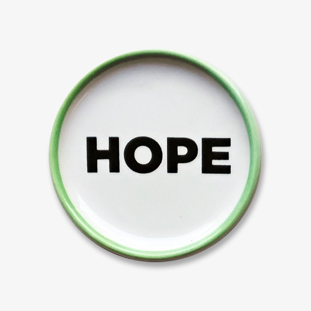 The Hope Trinket Dish is a handmade round white porcelain plate with a green rim and the word HOPE in bold black letters at the center, inspired by the Obama Foundation.
