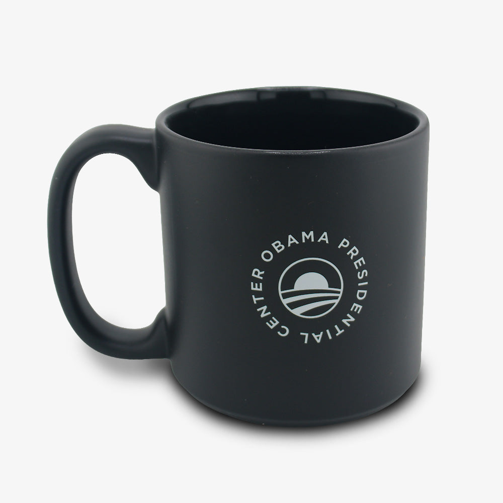 The Obama Presidential Center Script Mug is a matte black ceramic mug featuring a white circular logo with a stylized rising sun, shown against a clean white background.
