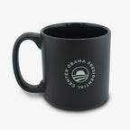 The Obama Presidential Center Script Mug is a matte black ceramic mug featuring a white circular logo with a stylized rising sun, shown against a clean white background.
