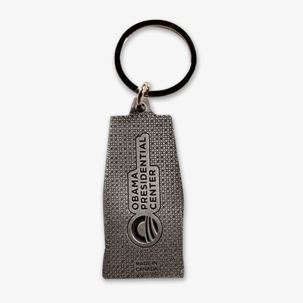 The Obama Presidential Center Building Keychain is a silver-tone keychain with a textured rectangular tag and round keyring, engraved with "Made in Canada," making it a distinguished presidential keepsake.