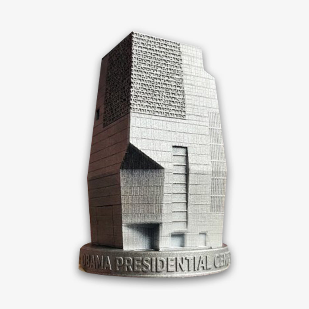The Obama Presidential Center Building Figurine is a gray, geometric architectural model with textured surfaces and a labeled circular base—a distinctive collectible keepsake.