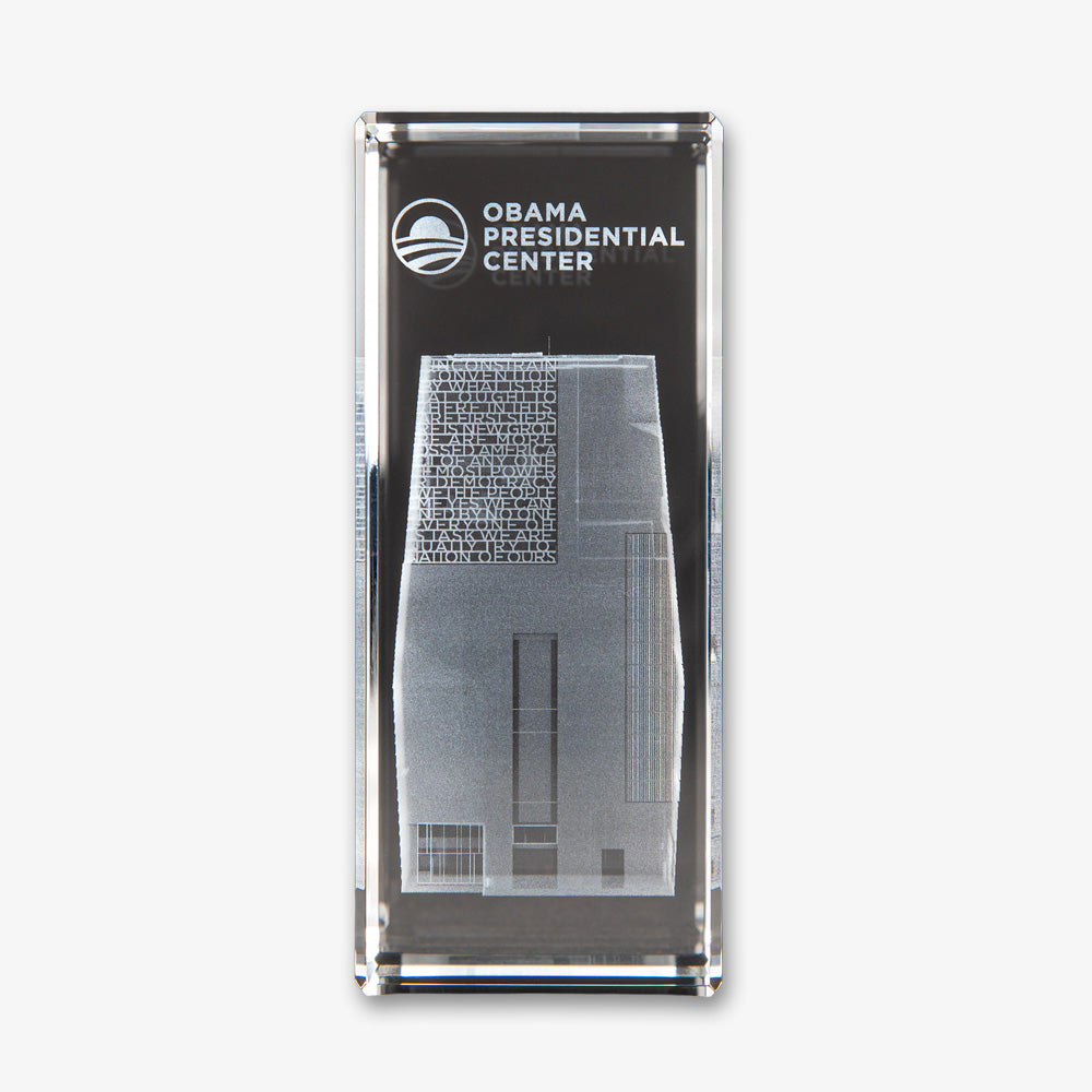 The Obama Presidential Center Building Crystal Cube is a clear rectangular crystal featuring an etched image of the building, the center’s name, and a circular logo above. The product stands out beautifully against a white background.