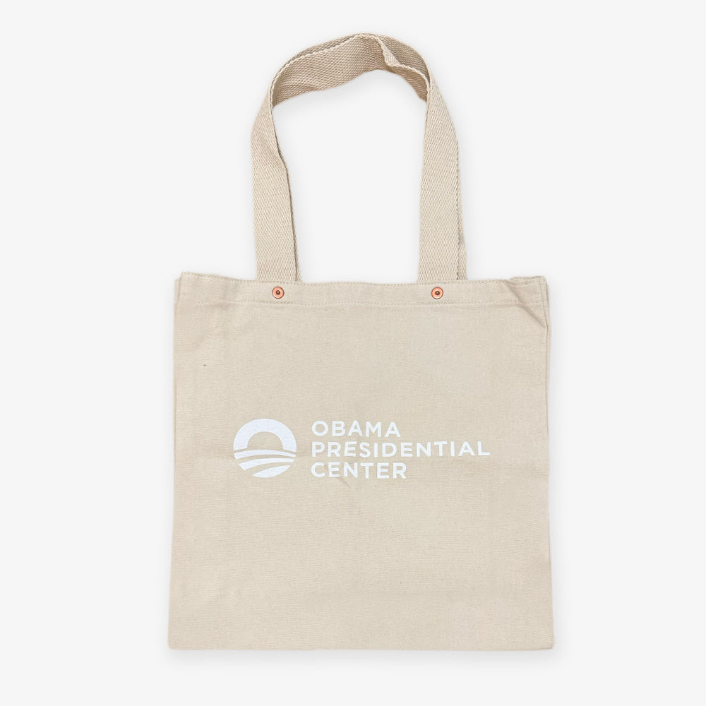 The Obama Presidential Center "You Are America" Speech Tote is a beige canvas bag with two handles, featuring the Obama Foundation tote logo and text in white on the front against a plain white background.
