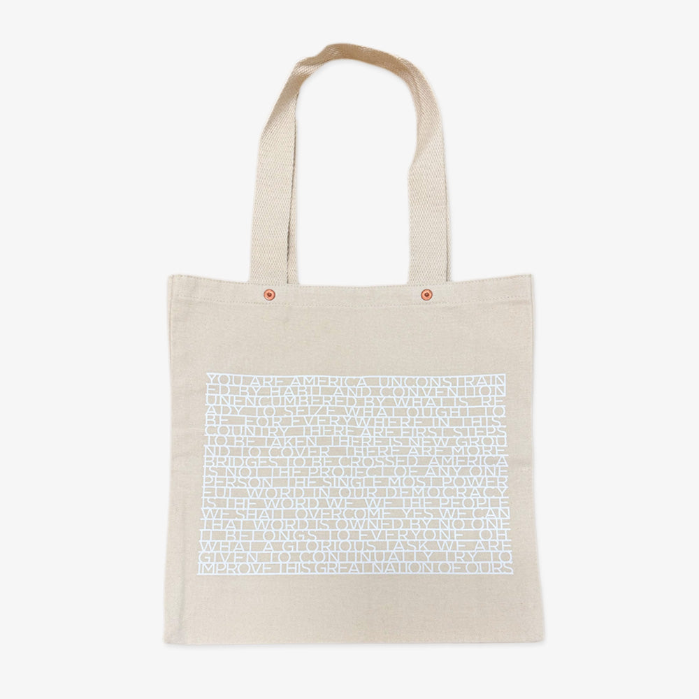 The Obama Presidential Center "You Are America" Speech Tote is a beige canvas bag with two handles and a block of dense white uppercase text covering most of the front.