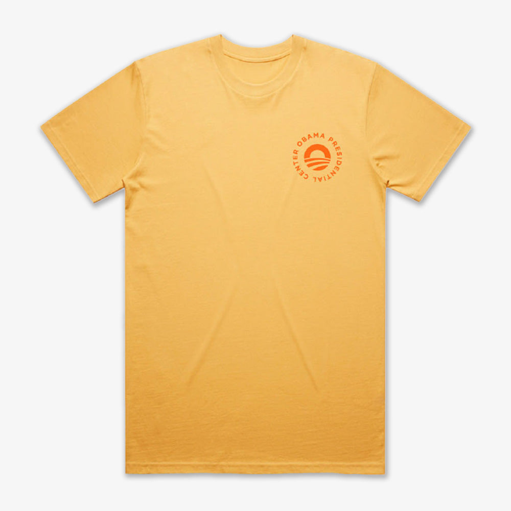 The Obama Presidential Center Hope to Action T-shirt - Sunset is a plain yellow tee with a small orange circular logo and text on the left chest, laid flat on a white background. Simple yet inspirational apparel.