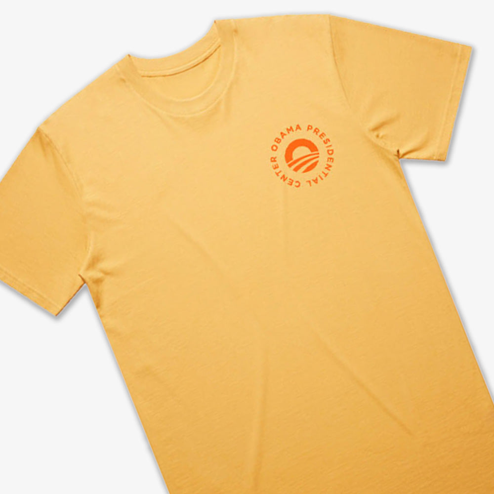 The Obama Presidential Center Hope to Action T-shirt - Sunset is a light orange tee with a small circular "Obama Presidential Center" logo and stylized O on the chest, shown laid flat on a white background.