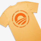 The Obama Presidential Center Hope to Action T-shirt - Sunset features a light orange color and a bold sun-and-waves design on the back, encircled by the words RESILIENCE, HOPE, COMMUNITY, IMAGINATION, EMPATHY, and COURAGE.