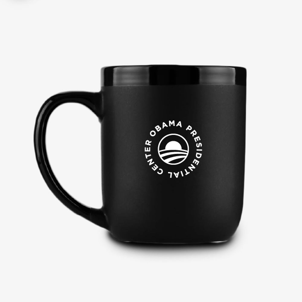 The Obama Presidential Center Script Mug is a matte black ceramic mug with a handle, showcasing the Obama Presidential Center logo and text in white on the front. Placed against a plain white background, this official Obama Foundation mug makes a sleek and meaningful addition to your collection.