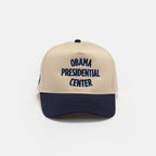 The Obama Presidential Center Home Court Cap, in beige and navy blue with "Obama Presidential Center" embroidered in dark blue on the front, is featured as official Obama Foundation merchandise against a plain white background.