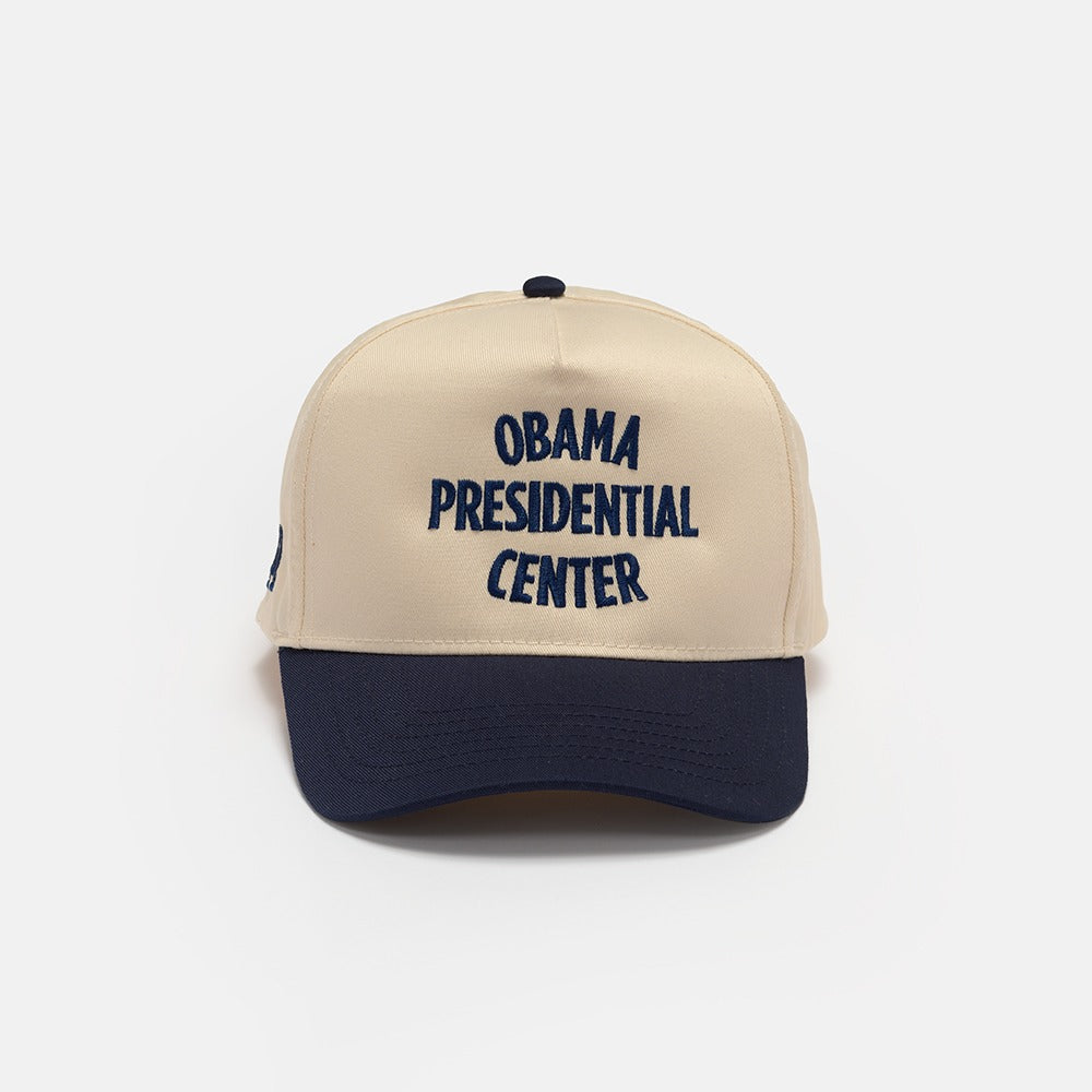 The Obama Presidential Center Home Court Cap, in beige and navy blue with "Obama Presidential Center" embroidered in dark blue on the front, is featured as official Obama Foundation merchandise against a plain white background.