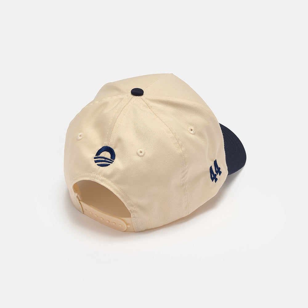 The Obama Presidential Center Home Court Cap in beige features a dark blue curved logo above the adjustable strap, the number 44 embroidered on the side, and a navy brim—official merchandise of the Obama Foundation.