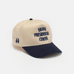 The Obama Presidential Center Home Court Cap is beige and navy with "OBAMA PRESIDENTIAL CENTER" on the front and "44" on the side. This official Obama Foundation cap is shown against a plain white background.