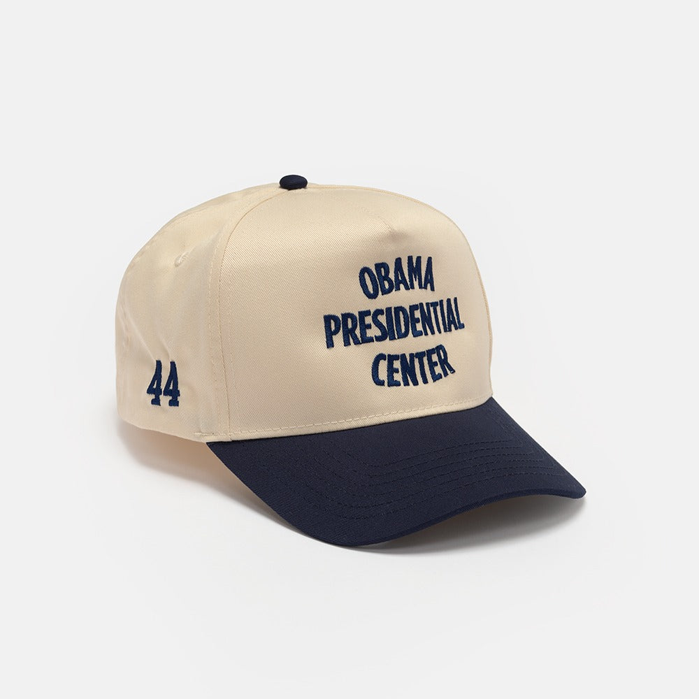 The Obama Presidential Center Home Court Cap is beige and navy with "OBAMA PRESIDENTIAL CENTER" on the front and "44" on the side. This official Obama Foundation cap is shown against a plain white background.