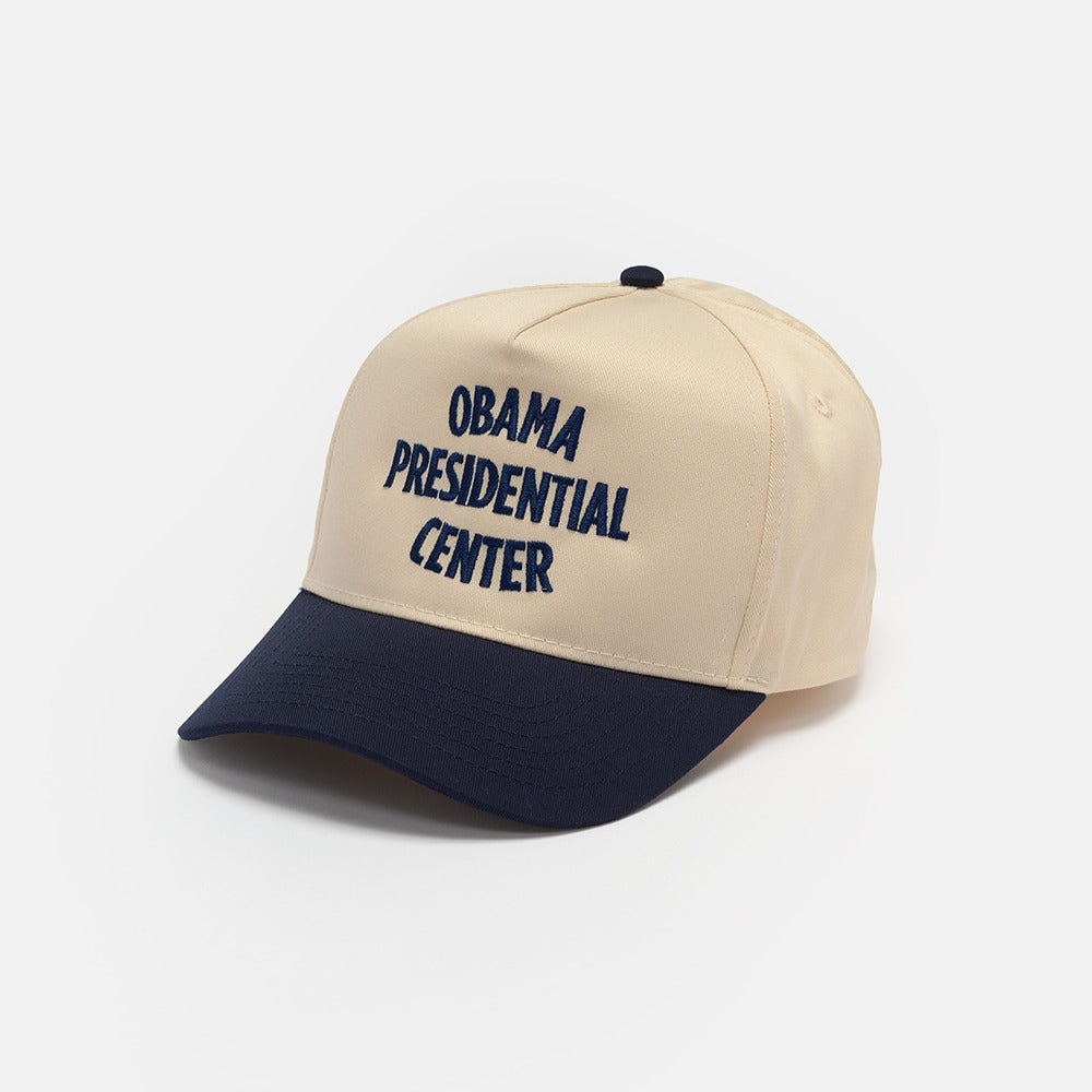 The Obama Presidential Center Home Court Cap is a beige hat with a navy blue brim and “OBAMA PRESIDENTIAL CENTER” embroidered in navy blue—an official piece of Obama Foundation merchandise.