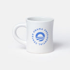 The Obama Presidential Center Logo Mug is a white ceramic mug featuring a blue circular logo with "Obama Presidential Center" text around a stylized O symbol, shown on a plain light gray background.