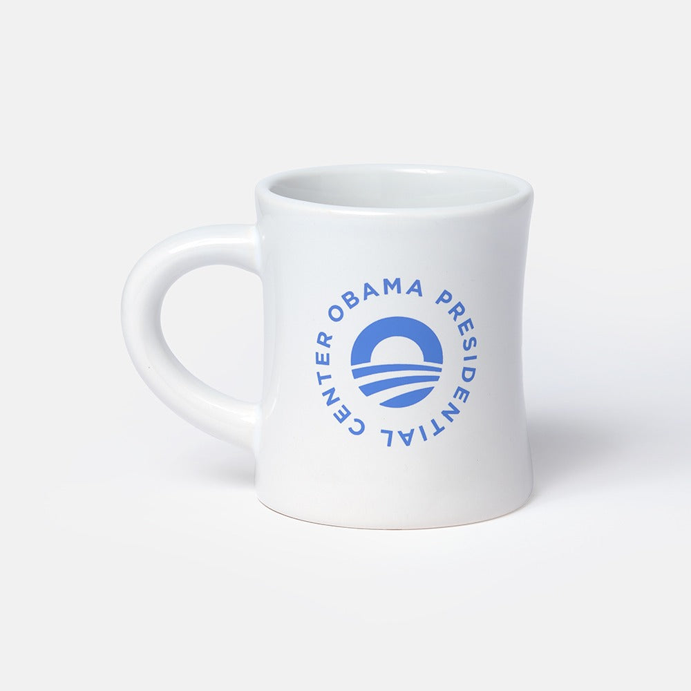 The Obama Presidential Center Logo Mug is a white ceramic mug featuring a blue circular logo with "Obama Presidential Center" text around a stylized O symbol, shown on a plain light gray background.