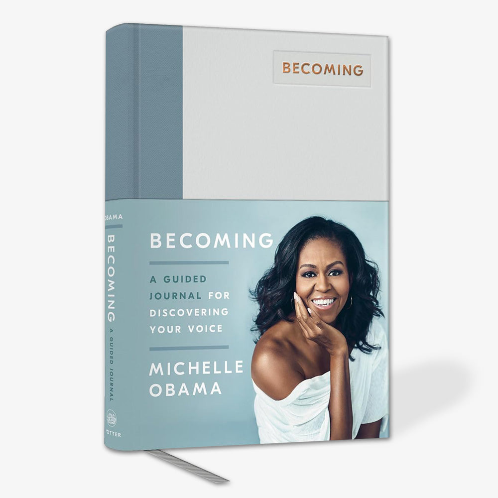 Becoming: A Guided Journal for Discovering Your Voice is an inspirational self-discovery journal by Michelle Obama, featuring a smiling cover photo of her seated on light blue and white, with a silver ribbon bookmark.