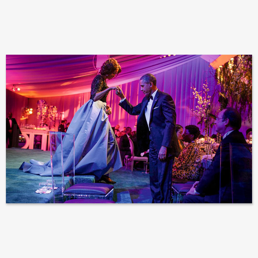 At an elegant, purple-lit event, a man in a tuxedo helps a woman in a formal gown onto a platform—an atmosphere reflecting the style and grace celebrated in The Look, Michelle Obama—with flowers and seated guests in the background.