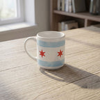 The Chicago Star Mug—a white mug with two red stars and light blue stripes inspired by the Chicago flag—rests on a wooden table. Proceeds benefit the Obama Foundation. Shelves and a window can be seen in the background.