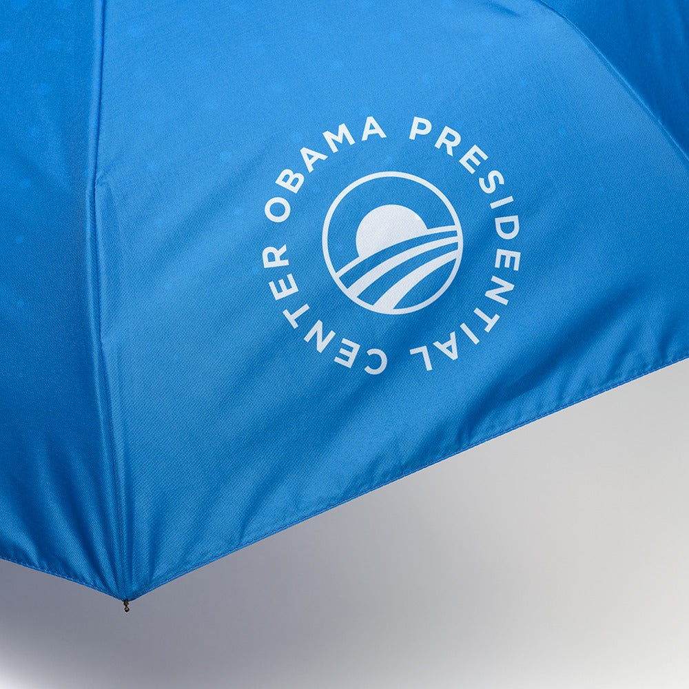 The Obama Presidential Center Umbrella is blue with the Obama presidential logo and "Center Obama Presidential" printed in a white circular design, inspired by Chicago’s South Side and supported by the Obama Foundation.