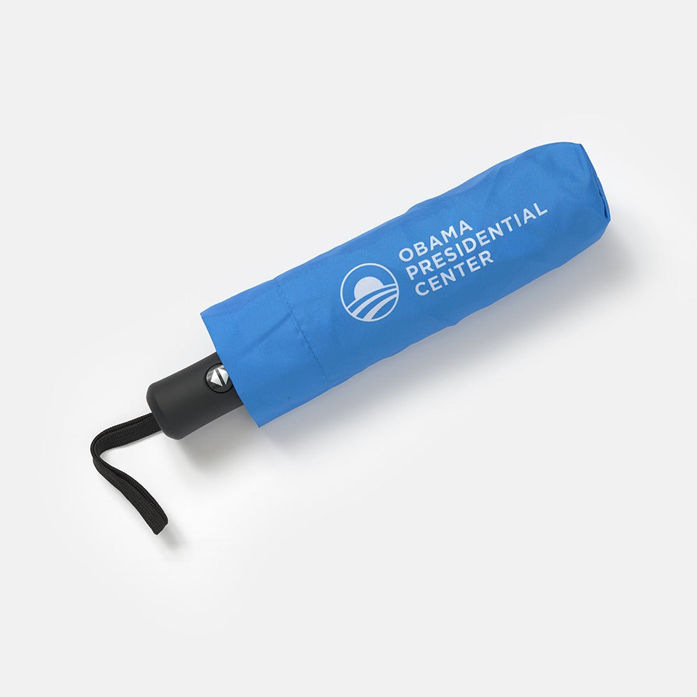 The Obama Presidential Center Umbrella, in blue with logo and text and a matching sleeve, rests on a light gray background—ideal for supporters of the Obama Foundation and Chicago's South Side.