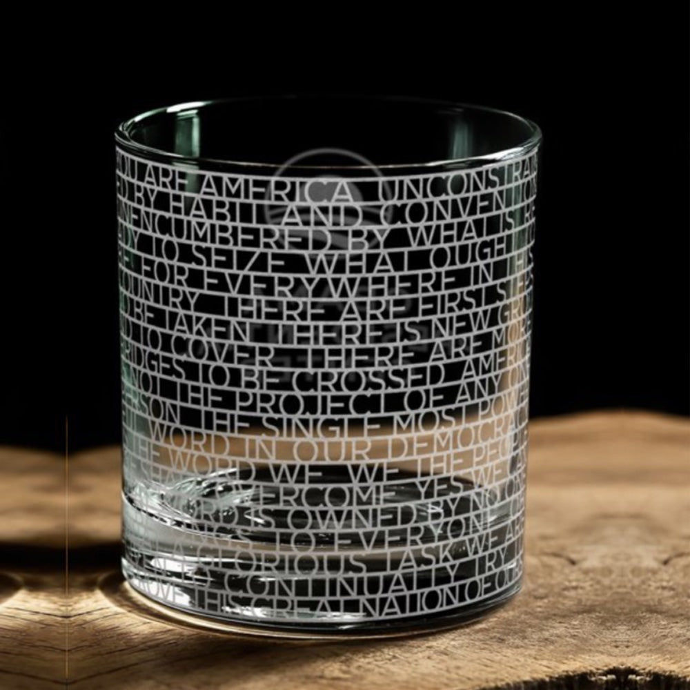 The Obama Presidential Center "You Are America" Speech Rocks Glass rests on a wooden surface, featuring tightly etched uppercase text from the speech wrapped around its clear glass. The words form a striking design against a black background.