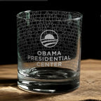 The Obama Presidential Center "You Are America" Speech Rocks Glass, featuring the center's logo and text, is shown on a wooden surface with a dark background.