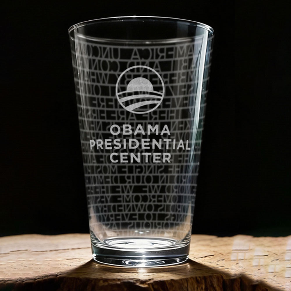 The Obama Presidential Center "You Are America" Speech Pint Glass, featuring the center’s logo and speech text, is exclusive Obama Foundation merchandise displayed on wood with a dark background.