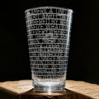 The Obama Presidential Center "You Are America" Speech Pint Glass features bold, etched uppercase text in horizontal bands. Displayed on a wooden surface, the white lettering stands out as a unique Obama Foundation collectible.