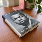 The hardcover book John Lewis, A Life rests on a wooden table by a window, with a green potted plant and a pink patterned object in the background.
