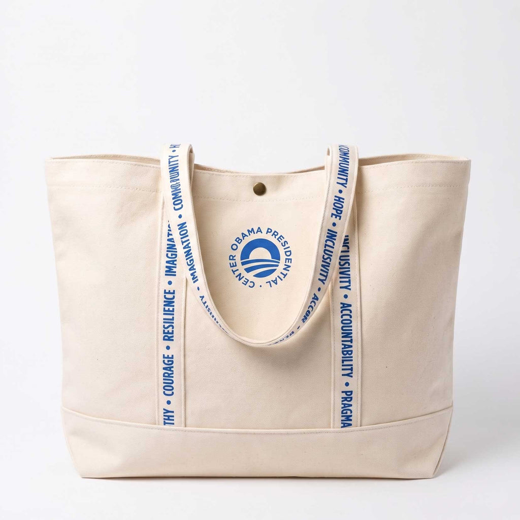 The Obama Presidential Center Logo Tote is a cream canvas bag with two handles printed “Courage” and “Resilience.” It features a blue circle logo and "Obama Presidential Center" text—stylish and inspiring from the Obama Foundation.