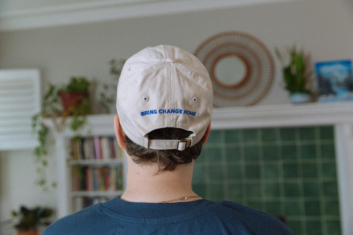 Someone wears the Obama Presidential Center Symbol Cap—an off-white, unisex cotton hat with “BRING CHANGE HOME” on the back—standing indoors amid green plants, a bookshelf, and a round decorative mirror.