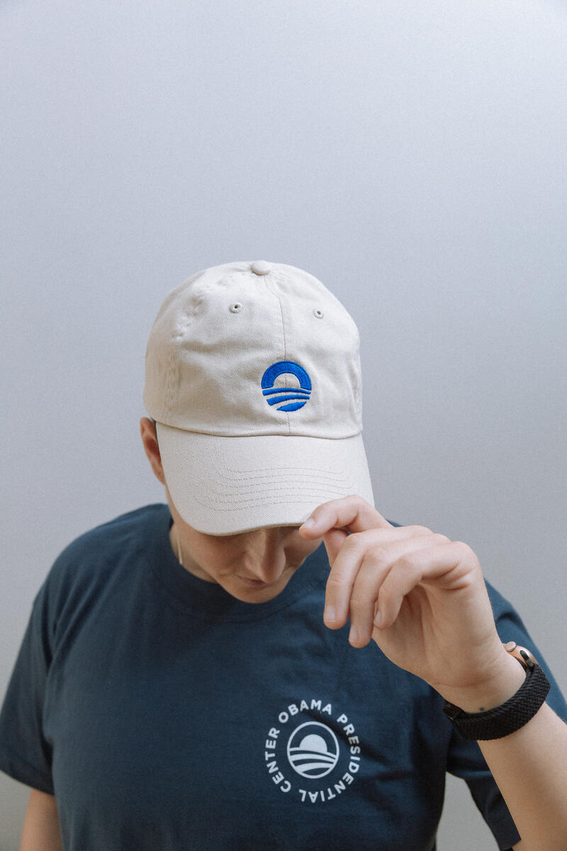 Wearing the Obama Presidential Center Symbol Cap and a navy blue T-shirt, both with the center’s logo, a person stands against a light gray background, tilting their head down to touch the cap's brim.