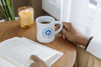A person sits at a round wooden table by a curtained window, reading a book and holding the Obama Presidential Center Logo Mug. Nearby, a lit candle and green plant complete the cozy scene.