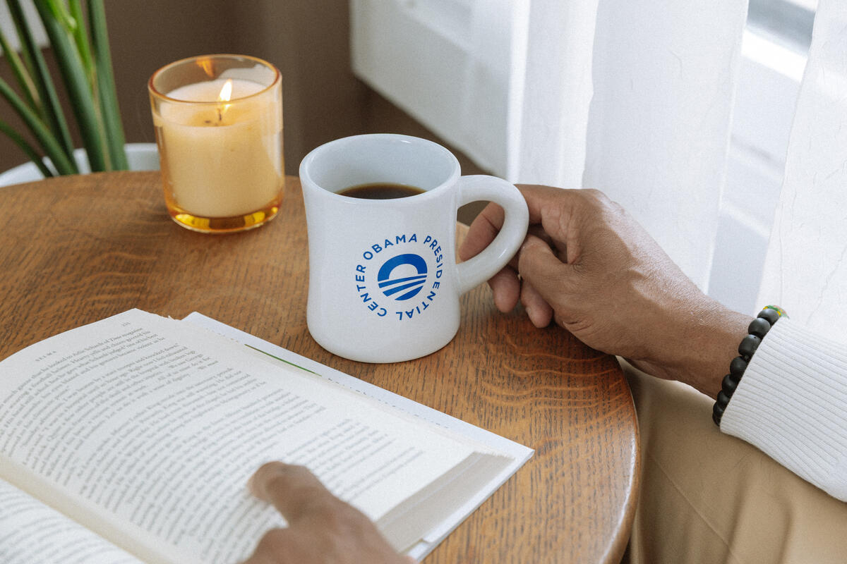A person sits at a round wooden table by a curtained window, reading a book and holding the Obama Presidential Center Logo Mug. Nearby, a lit candle and green plant complete the cozy scene.