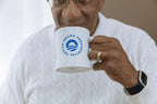 A person in a white sweater and watch drinks from an Obama Presidential Center Logo Mug, partially covering their face with the white ceramic mug featuring the center’s logo and text.