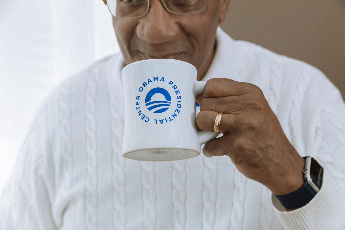A person in a white sweater and watch drinks from an Obama Presidential Center Logo Mug, partially covering their face with the white ceramic mug featuring the center’s logo and text.